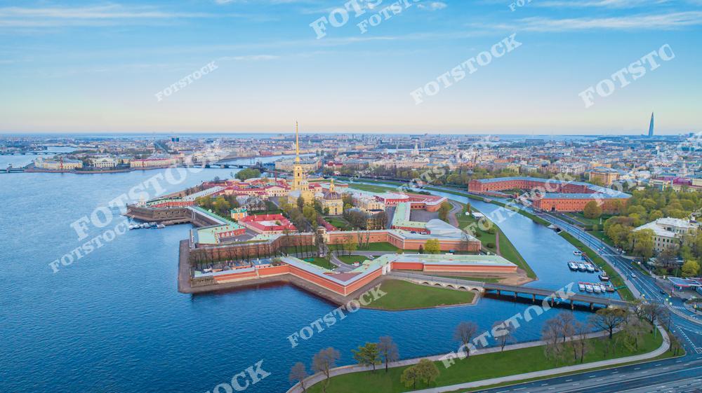 На фотографии изображена Петропавловская крепость — историческое сердце Санкт-Петербурга, расположенное на Заячьем острове. В центре кадра выделяется Петропавловский собор с золотым шпилем — одна из архитектурных доминант города. Вдоль крепостных стен протекает Нева и Кронверкский пролив. На горизонте виден небоскрёб Лахта Центра, а на переднем плане — набережные с молодой весенней зеленью. Вид с воздуха охватывает значительную часть центрального Петербурга.
